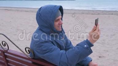 男子博主穿着蓝色羽绒服坐在沙滩的长椅上，在手机上聊天视频..